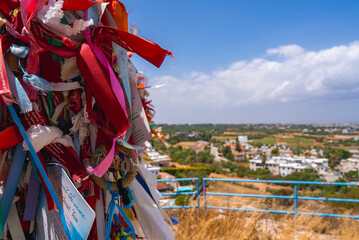 Vibrant ribbons tied to a structure with a rural Cyprus backdrop, featuring white buildings, green fields, dry grass, and a clear blue sky.