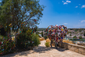 A tree adorned with vibrant ribbons and fabric strips stands on a stone paved pathway, surrounded by greenery and overlooking a residential area in Cyprus. © Aerial Film Studio