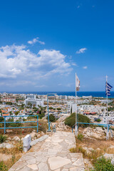 View of a coastal town in Cyprus with whitewashed buildings, a luxury hotel, greenery, and a stone pathway leading to a viewpoint with flags. © Aerial Film Studio