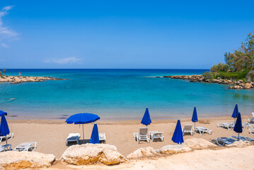 A tranquil beach in Cyprus featuring golden sand, turquoise waters, blue umbrellas, white lounge chairs, rocky outcrops, and lush greenery.