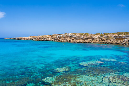Crystal clear turquoise waters with visible underwater rocks meet a rugged, rocky shoreline under a bright blue sky in a serene Cyprus coastal view.