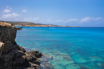 Crystal clear turquoise waters meet rugged rocky cliffs in Cyprus. The horizon shows sandy beaches and a distant resort town under a bright blue sky.