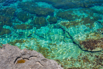 Clear turquoise waters reveal rocky formations beneath the surface, with a rugged rock edge in the foreground along the Cyprus coastline.