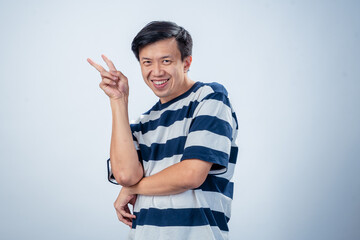 Asian man in a striped shirt making a secretive expression while raising a clenched fist near his mouth, as if plotting or thinking deeply, against a plain studio background