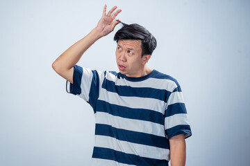 Asian man in a striped shirt lifts a strand of his hair while looking upward in confusion or curiosity, standing against a plain light background