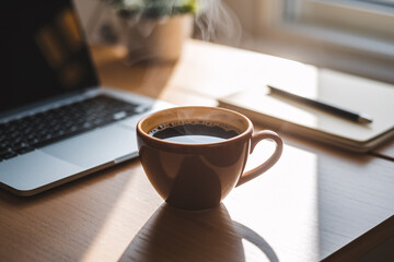 Cup of steaming coffee on desk. Cozy workspace with notebook in background