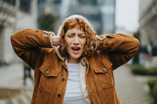 A woman in a brown coat covers her ears, seemingly distressed. The background depicts an urban cityscape, suggesting concepts like noise pollution, frustration, and sensitivity.