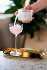 A hand pours a white liquid into a cocktail glass filled with pink cotton candy, placed on a golden tray on a speckled countertop. The scene captures a stylish drink preparation with a luxurious touch