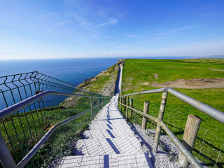A stairway descends along a cliffside path bordered by green fields and overlooking a vast blue sea