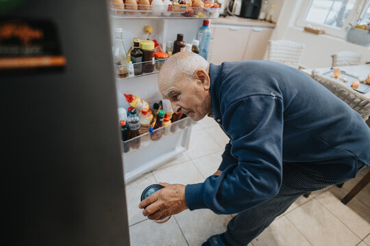 A senior man inspects a can of food in his kitchen while keeping the refrigerator open, showcasing a moment of daily life and home activity, emphasizing practicality and routine.