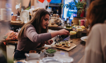 A woman arranging small appetizers on a wooden counter in a warmly lit bar. Various ingredients, utensils, and decorations create a warm and inviting atmosphere.