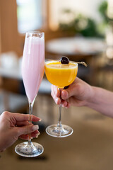 Two hands holding glasses of vibrant cocktails—one pink, one orange—garnished with cherries. A blurred background suggests a lively, celebratory atmosphere