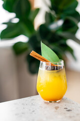 A vibrant yellow-orange cocktail with ice, garnished with a cinnamon stick and a green leaf, placed on a marble surface with a blurred green plant background, evoking freshness and summer vibes