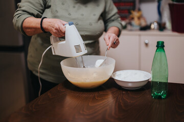 An elderly woman skillfully uses a hand mixer to prepare batter for baking. The scene captures a homey moment during a culinary activity.