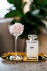 Elegant bottle labeled "LOVE IS... MADE FOR YOU" placed on a golden tray with a glass of pink cotton candy. Soft, romantic composition with a blurred green background, exuding luxury and charm