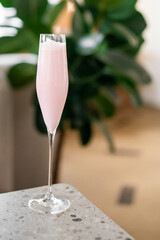 A tall, elegant glass filled with a pink, frothy drink sits on a speckled countertop. A blurred green plant in the background adds a stylish and refreshing touch