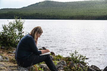 A woman is reading a book while sitting at the shore of a lake on a bright summer day in Sweden. She is enjoying the peace and quiet of nature.
