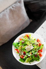 A vibrant salad with mixed greens, cherry tomatoes, olives, microgreens, and a seasoned cheese cube, served in a white bowl on a table, with a cozy couch in the background