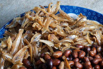 A close-up view of a traditional Malaysian side dish featuring crispy fried anchovies (ikan bilis) and roasted peanuts served on a blue plate. 