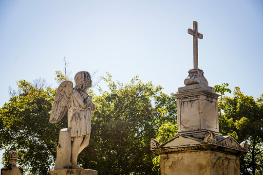 Statue of an angel before a cross in prayer. Blue sky and lush greenery surround it. S&atilde;o Jo&atilde;o Batista Cemetery, Rio de Janeiro, Brazil.