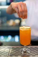 A bartender's hand sprinkles garnish onto a vibrant orange cocktail in a stemmed glass on a metal bar counter. The blurred background highlights the drink preparation in a professional bar setting
