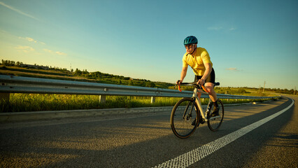 Fototapeta premium Man rides hard on wide road through nature terrain with intense expression against hills and soft sunset on background. Banner. Concept of sport, cycling, healthy lifestyle, endurance, nature.