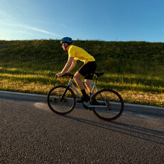 Fototapeta premium Rider in bright gear cycles near grassy terrain under clean blue sky with golden evening light and gentle sense of movement. Concept of sport, cycling, healthy lifestyle, endurance, nature.