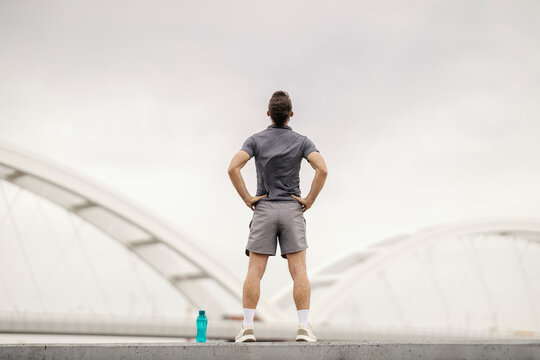 Back view of sportsman standing on riverbank with hands on hips and looking at river.