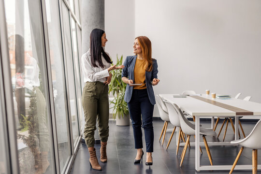 Two business casual businesswomen walking in office and discussing project.