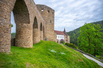 The Gothic Bridge of Velhartice Castle stands majestically over a green hillside, showcasing its stone arches. The surrounding landscape features trees and a pathway leading to the castle.