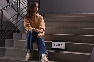 Cyber bullying. Upset teenage girl and laptop with text You Loser on stairs indoors. Space for text