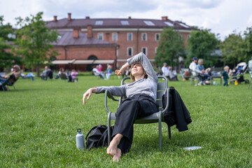 Woman lounging in park chair shielding eyes from sun, resting napping with water bottle on break after work. Urban green park relaxation, city break, relaxed female recharge, calm downtime in nature 