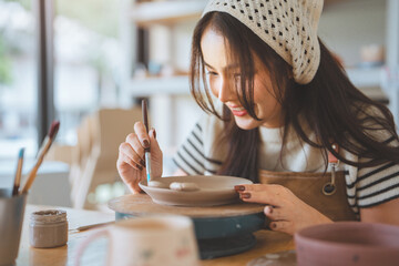 Asian woman artist painting handmade ceramic product in pottery studio – creative process of decorating sculpture with brush for small business, art workshop, and modern craft design startup