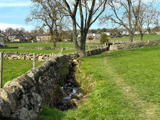 A narrow stream runs alongside a stone wall, with grassy fields stretching out under a clear blue sky. Bare trees dot the landscape, and houses are visible in the distance near, Guiseley, Leeds, UK