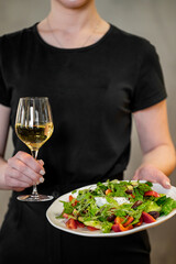 A person wearing a black shirt holds a glass of white wine and a plate with a fresh vegetable salad. The scene emphasizes healthy eating and a sophisticated dining experience in a minimalist setting