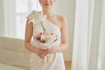 Caucasian young adult woman holding wedding bouquet with both hands standing indoors wearing elegant dress smiling softly wedding celebration
