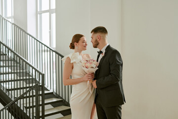 Caucasian young adult bride holding bouquet standing close to Caucasian young adult groom in formal suit near staircase during wedding celebration, both smiling at each other