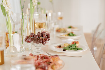 Wedding celebration table setting featuring elegant glassware, plates with appetizers, fresh grapes in glass bowl, floral arrangements, soft focus background, no people visible