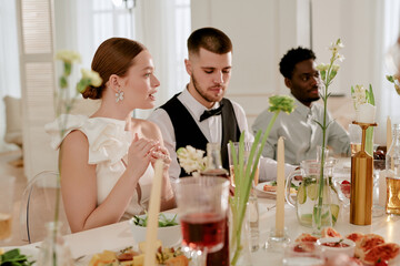 Caucasian young adult woman sitting beside Caucasian young adult man and Black young adult man at wedding celebration table, engaging in conversation and enjoying festive meal together