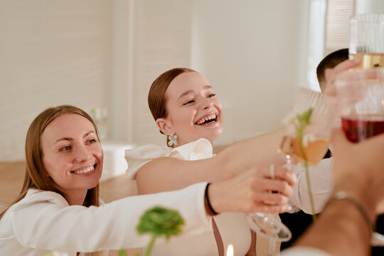 Caucasian young adult women smiling and raising glasses in celebratory toast during wedding celebration, surrounded by friends, joyful expressions visible, hands holding drinks extended