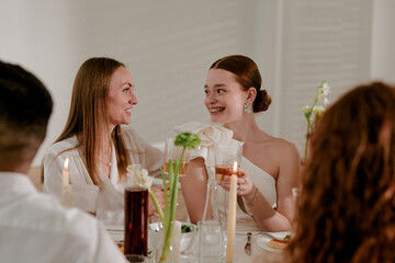 Caucasian young adult woman in wedding dress smiling and clinking glasses with Caucasian young adult woman during wedding celebration, surrounded by guests at decorated table