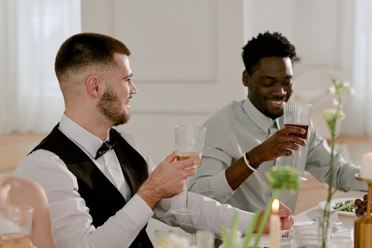Caucasian young adult man and Black young adult man sitting at wedding table raising glasses in toast smiling and interacting during wedding celebration