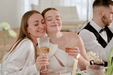 Caucasian young adult bride and Caucasian middle aged woman smiling and embracing while holding wine glasses at wedding celebration, Caucasian young adult man sitting beside them
