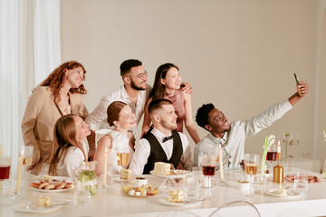 Group of young adult multiethnic men and women smiling and posing for selfie at wedding celebration, sitting and standing around decorated banquet table with food and drinks