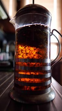 This footage conveys a warm and inviting mood, depicting tea leaves actively brewing in a French press, illuminated by soft backlight, ideal for illustrating cozy moments, a relaxing start to the day