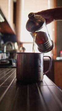 This footage conveys a comforting and authentic mood, depicting a hand pouring freshly brewed tea or coffee from a French press into a rustic metal mug, perfect for illustrating daily rituals
