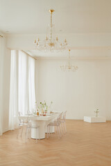 Empty wedding celebration venue featuring elegant table set for event with transparent chairs and minimalist floral arrangements, bright spacious room with chandeliers and parquet floor