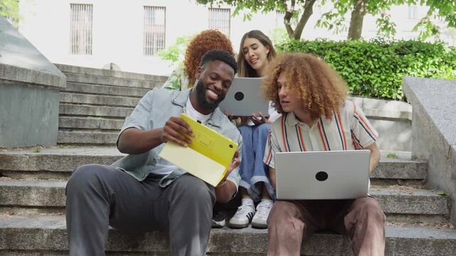 University students working together on academic project, sitting on outdoor steps, sharing ideas using digital devices with diverse team members
