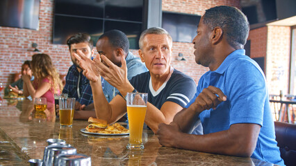 Multi Cultural Group Of Male Friends Watching Game In Sports Bar Eating Food