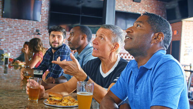 Multi Cultural Group Of Male Friends Watching Game In Sports Bar Eating Food